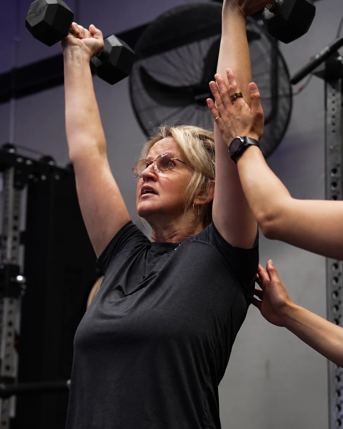 A member pressing two dumbbells overhead with hands on support from a coach at Injury Active Clinic